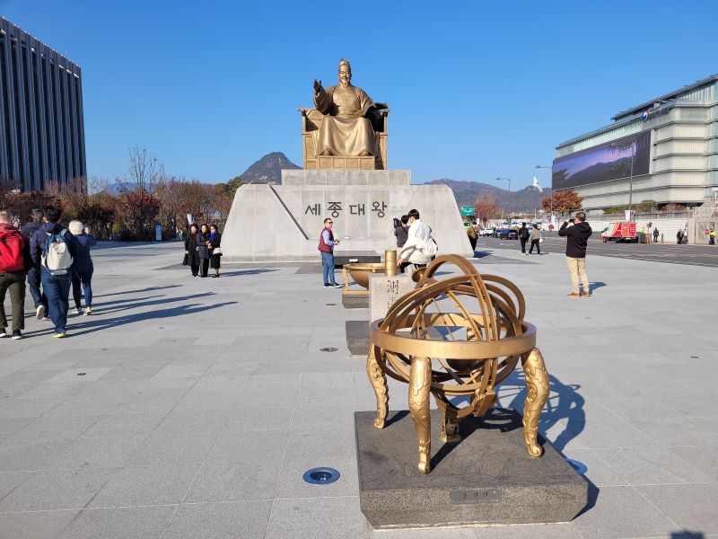 Pomnik króla Sejonga w Seulu, przed nim na planu ludzie, a na pierwszym planie rzeźba sfery armilarnej