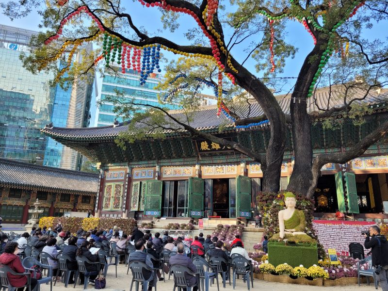 Świątynia buddyjska w Seulu z drzewem stojącym przed budynkiem, obwieszonym religijnymi strojeniami, przed budynkiem siedzą ludzie na krzesłach