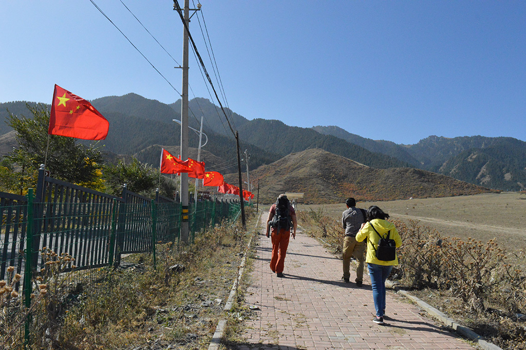 Troje naukowców idzie drogą w kierunku gór. Na ogrodzeniu z lewej strony drogi widoczne chińskie flagi // Three scientists walking towards the mountains. There are Chinese flags visible on the fences on the left