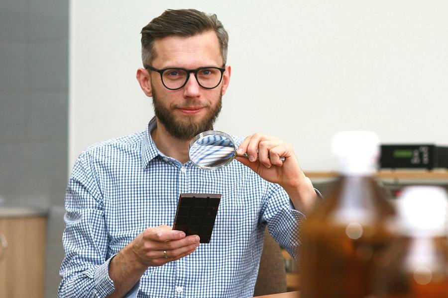 Dariusz+Kajewski_0 Profesor siedzi przy stole laboratoryjnym, trzymając w jednej dłoni lupę, a w drugiej prostokątny element przypominający panel lub próbkę materiału. Na pierwszym planie widoczne są rozmyte butelki laboratoryjne, a w tle znajduje się wyposażenie pracowni