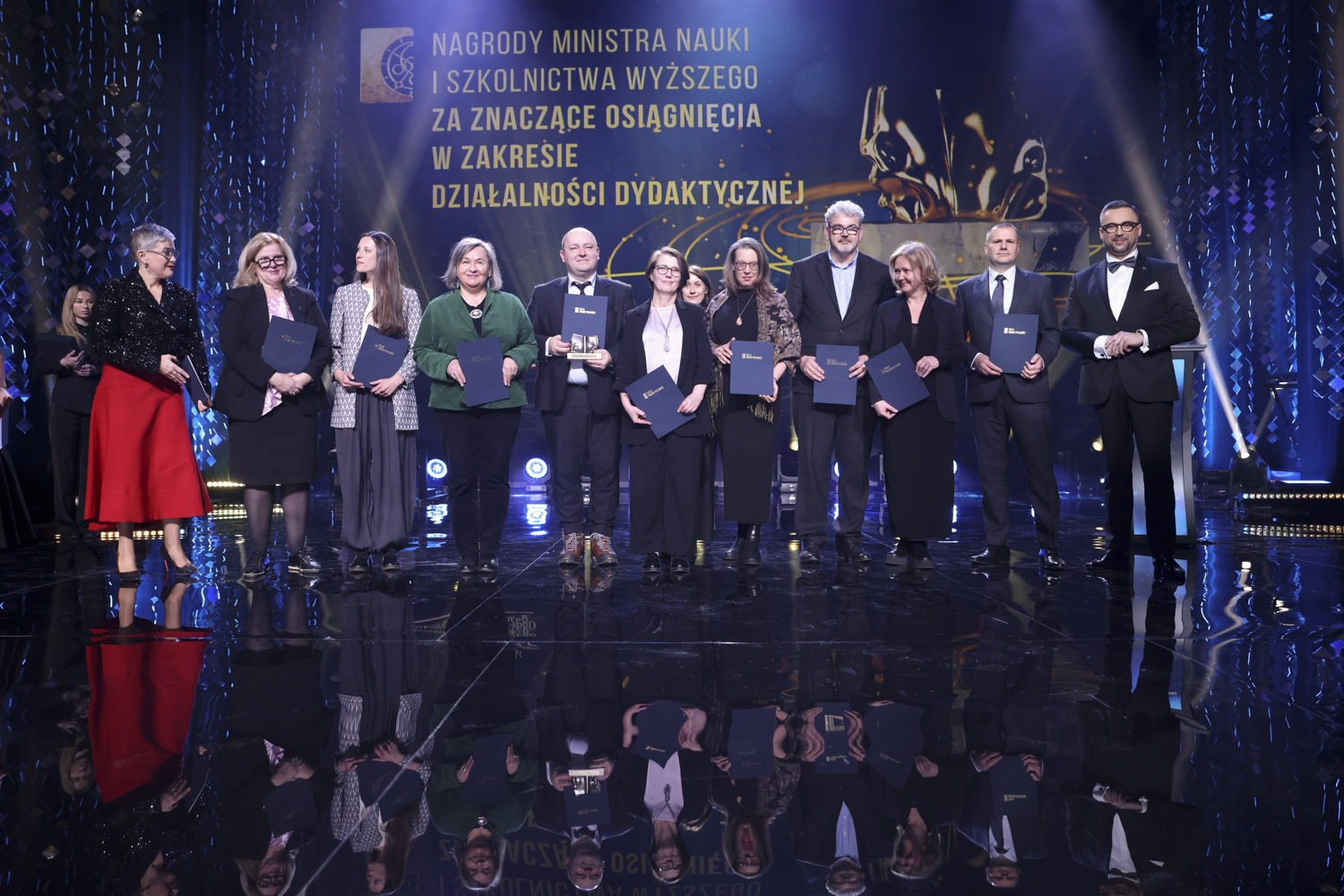 Gala Nauki Polskiej 2 Grupa osób stoi na scenie podczas uroczystości wręczenia nagród. Wszyscy trzymają w rękach granatowe teczki. Po lewej stronie widoczna jest osoba prowadząca wydarzenie w czerwonej spódnicy i ciemnej marynarce. W tle znajduje się duży ekran z napisem: ‘Nagrody Ministra Nauki i Szkolnictwa Wyższego za znaczące osiągnięcia w zakresie działalności dydaktycznej’. Scena jest oświetlona reflektorami, a dekoracja utrzymana w niebiesko‑złotej kolorystyce.