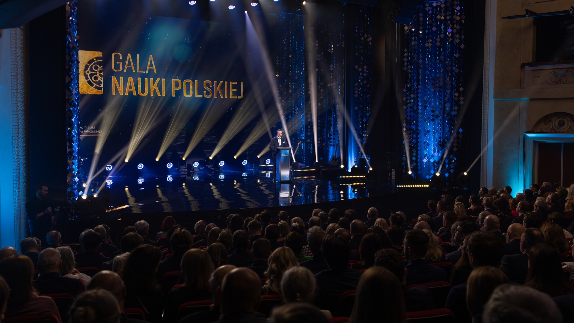 Gala nauki polskiej1 Widok dużej sali teatralnej wypełnionej publicznością. Na scenie, oświetlonej licznymi reflektorami, stoi osoba przemawiająca z mównicy. W tle widoczny jest ekran z napisem ‘Gala Nauki Polskiej’ oraz dekoracyjne, niebieskie elementy scenografii