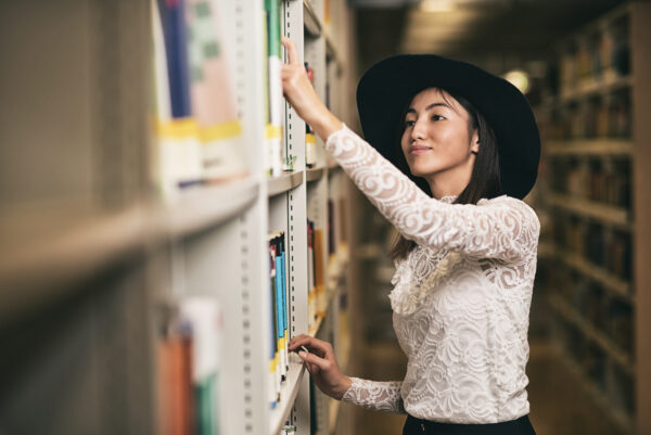 uśmiechnięta dziewczyna w kapeluszu wybierająca książki w bibliotece