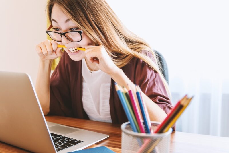 Młoda kobieta w okularach zagryzająca w zębach ołówek patrzy na monitor laptopa, obok stoi kubek z ołówkami