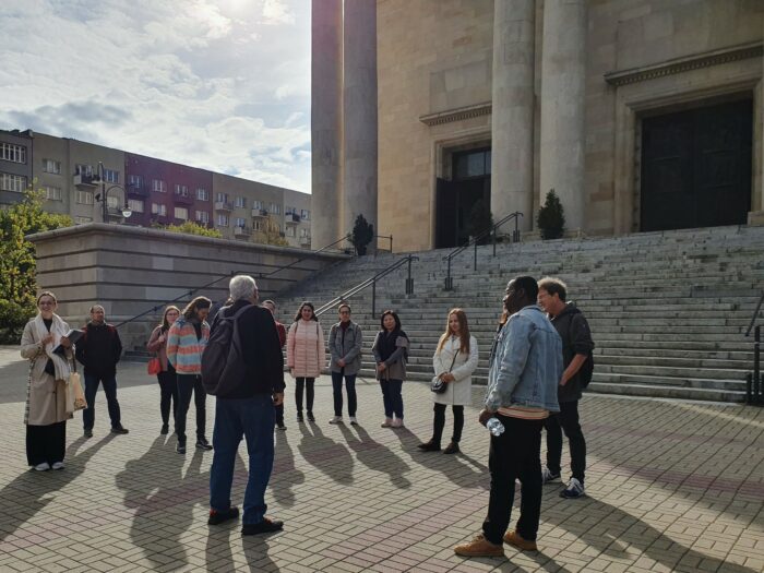 A group of people standing in front of the cathedral, in a circle, and listening to a guide. Grupa ludzi stojąca w okręgu przed katedrą i słuchająca przewodnika.
