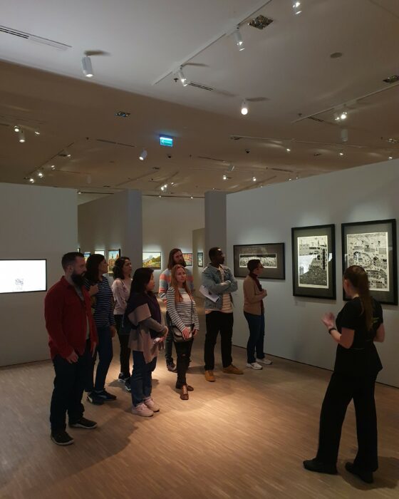 Group of people seeing an exhibition with paintings, listening to the museum guide. Grupa ludzi zwiedzająca wystawę obrazów z przewodniczką muzeum.