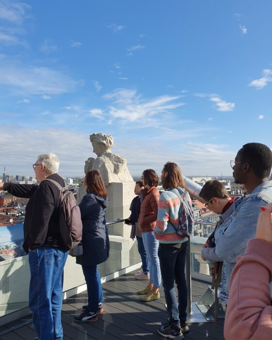 People looking at the city from the roof with the guide pointing to the buildings. Grupa ludzi podziwiająca panoramę miasta z góry, z przewodnikiem wskazującym zabudowania.