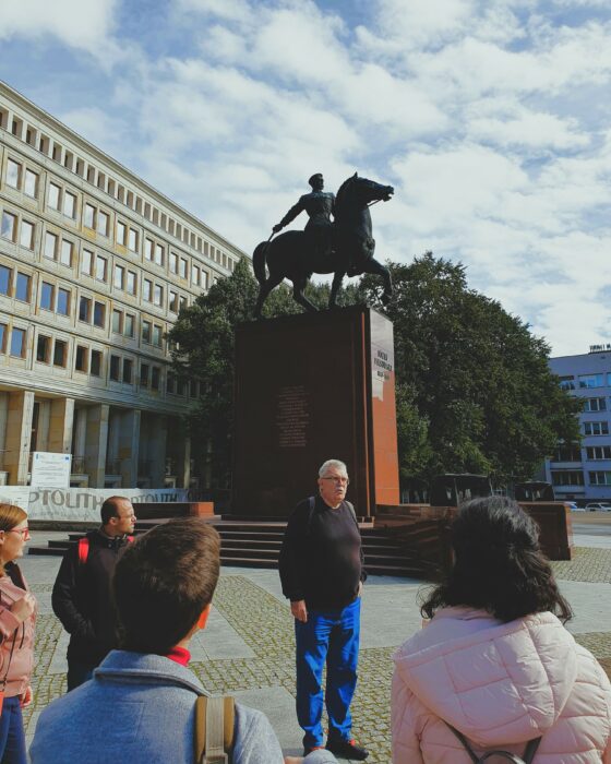 A group of people listening to the guide and looking at the monument of a general on the horse. Grupa osób słuchająca przewodnika przed pomnikiem generała na koniu.