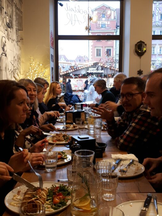 A group of people sitting on the both sides of the table and eating dinner. The window comming out to the chrismas market in the background. Grupa ludzi siedząca przy stole i jedząca obiad. W tle przez okno widać budkę jarmarku świątecznego.