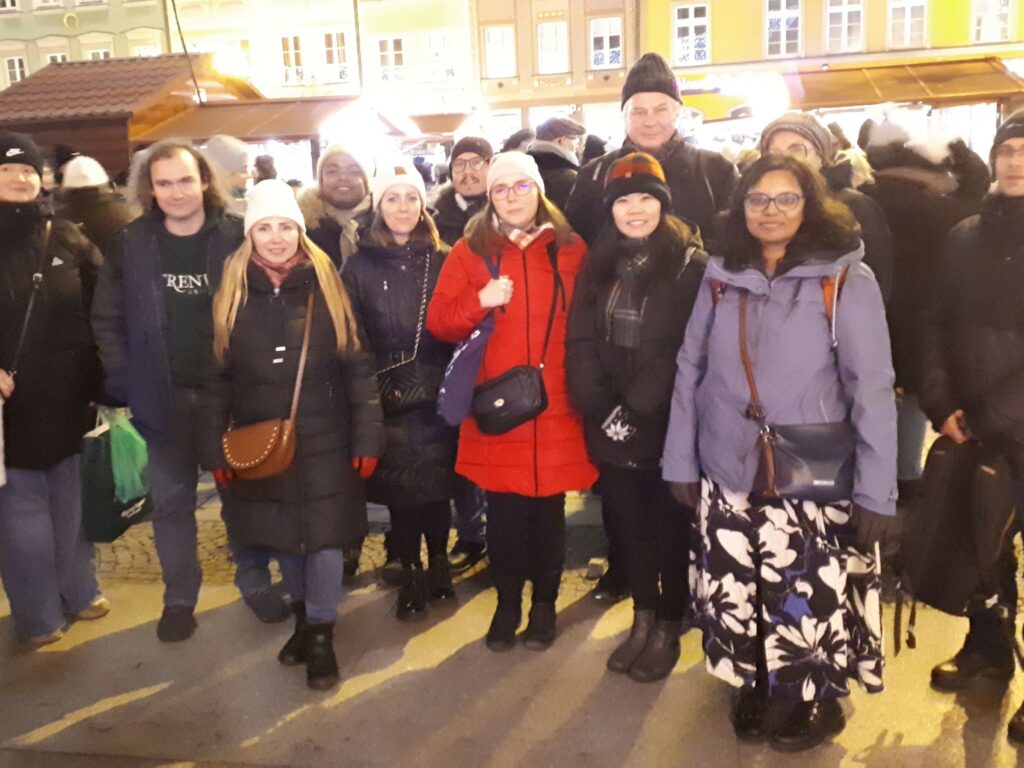 A group of people posing for a photo in the evening with roofs of Christmas Market behind them. Grupa uczestników wycieczki pozująca do zdjęcia wieczorem, z dachami świątecznych straganów w tle.