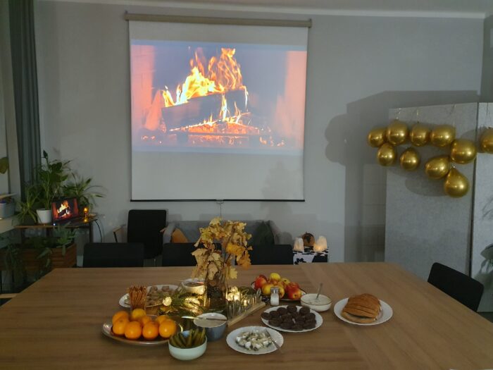 View of the room with decorated table an which there is some food, lights and a buquet of dry yellow leaves. Screen with a fireplace and golden baloons in the background. Udekorowany pokój ze stołem pośrodku. Na stole widać potrawy, światełka i bukiet suchych żótych liści. W tle ekran z widokiem ognia palącego się w kominku i wiszące złote balony.