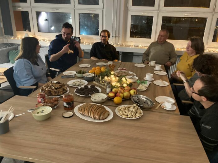 A group of six people sitting at the table laid with food listening to a man explaining something. Grupa sześciu osób siedząca przy zastawionym stole i słuchająca mężczyzny, który o czymś opowiada.