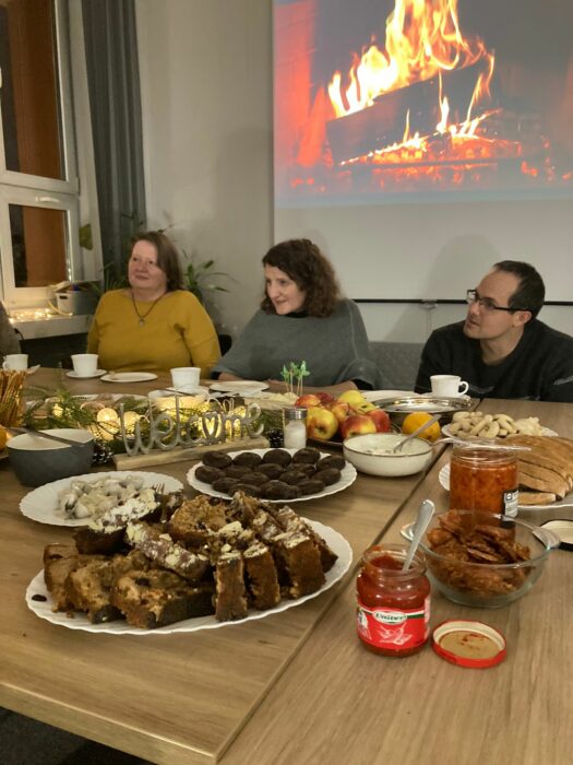 Three people: two women and one man sitting at the table listening to someone on the other side of the table. The view of a burining fireplace behind them. Dwie kobiety i mężczyzna siedzący przy zastawionym stole i słuchający kogoś, kto siedzi po drugiej stronie stołu. W tle widok na wyświetlony kominek z palącym się polanem.