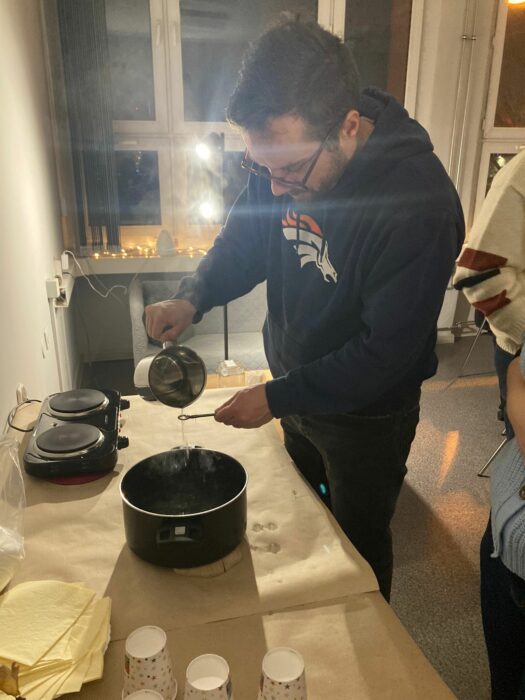 A man standing near the table and laying hot wax from the one pot through the keyhole to the other pot with cold water. Mężczyzna stojący przy stole i lejący gorący wosk przez dziurkę od klucza do garnka wypełnionego zimną wodą.