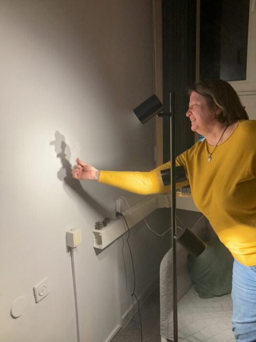 A lady in yellow sweater putting the wax shape to the wall lit by the lamp so that she can see its shadow of the shape she holds. Kobieta w żółtym swetrze przykładająca do ściany odlew z wosku, żeby zobaczyć cień kształtu na oświetlonej ścianie.