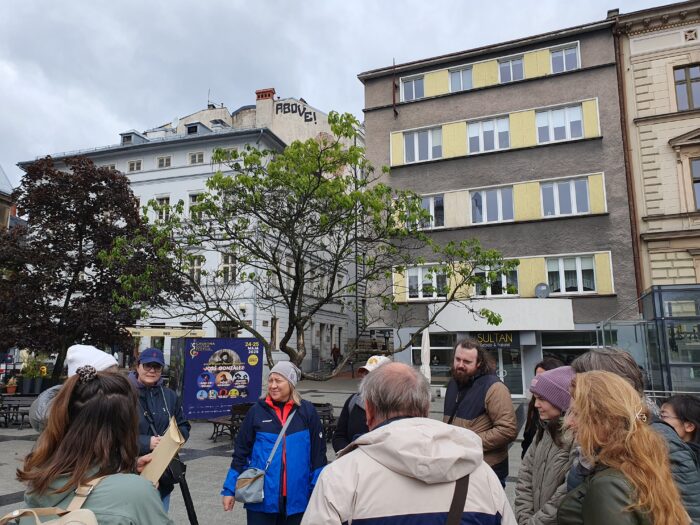 A group of people standing in the city listening to a guide. Grupa ludzi stojących w centrum Bielska i słuchająca instrukcji koordynatorki gry.