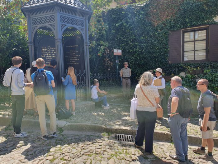 A group of people listetning o the guide, standing next to the towe-like monument. Grupa ludzi słuchająca przewodnika stojąca obok studni obudowanej na kształt wieży.