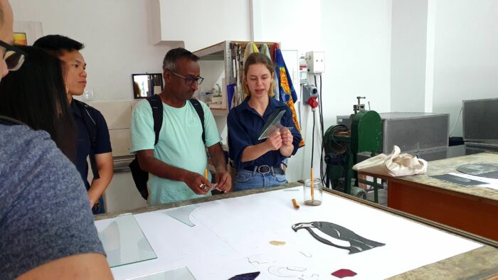 Small group of people in the workshop looking at the woman holding a pane of glass. Niewielka grupa w pracowni patrząca na kobietę rozdzielającą dwa kawałki szyby.