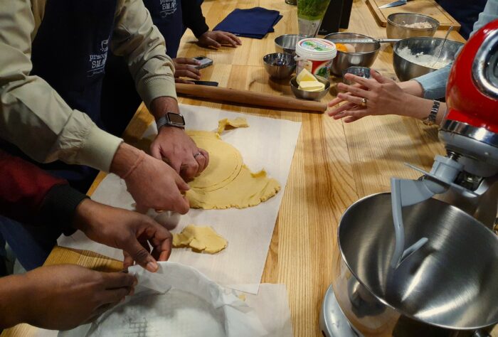Four pair of hands preparing the dough for the cake with cooking equipment around. Cztery pary rąk przygotowujące ciasto w otoczeniu przyborów kuchennych.