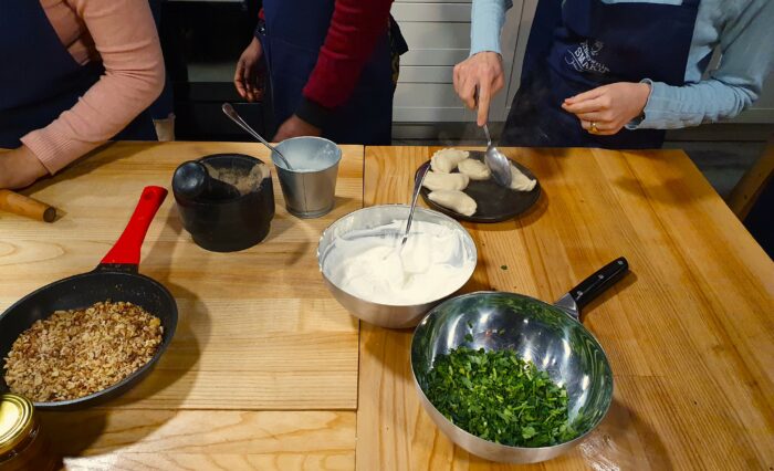 Hands serving piegori, a bowl of yogurt, with a bowl of parsley and a pan with nuts on the table beside them. Ręce serwujące pierogi, obok, na stole miska z jogurtem, miska z pietruszką oraz patelnia z orzechami.