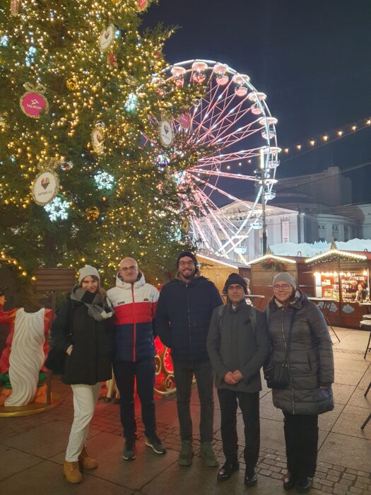 Five people posing for a photograph in the evening. A decorated Christmass tree and ferris wheel with lights in the background. Pięć osób pozujących do zdjęcia. W tle rozświetlona choinka i świecąca karuzela - młyńskie koło.