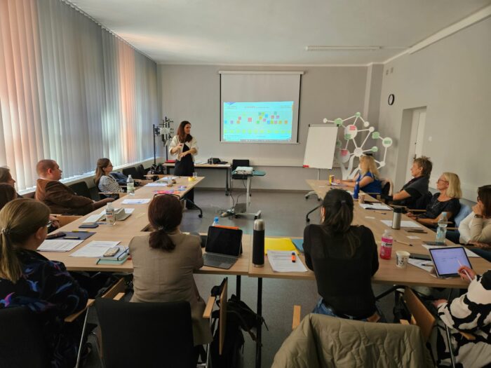 Group of people listetning and the trainer sitting in the room and looking at the screen. Grupa uczestników szkolenia oraz trenerka poldczas szkolenia patrzący na ekran z wyświetloną prezentacją.