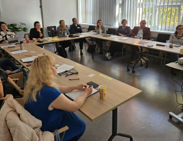 Group of training participants sitting at the table and listening to the trainer. Grupa uczestników szkolenia siedząca przy stołach ułożonych w kształcie podkowy.