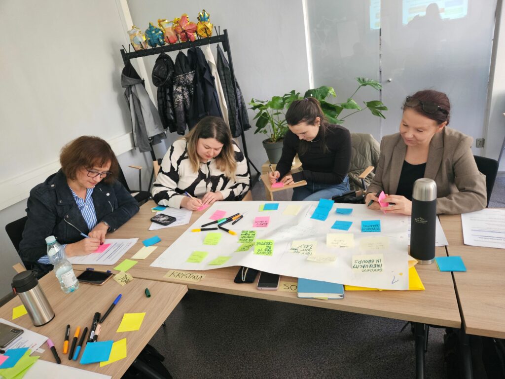 Four women writing at the post-it notes and sticking them at the white sheet of paper. Cztery kobiety siedzące przy stole i piszące na kolorowych karteczkach, które przyklejają do białego arkusza brystolu.