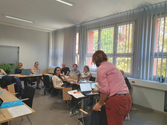 Group of training participants looking at the trainer, who prepares the presentation on his computer with his back to the photographer. Grupa uczestników szkolenia patrząca na trenera, który przygotowuje prezentację na komputerze, obrócony tyłem do fotografa.