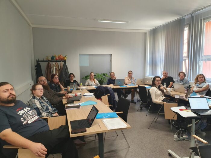 Group of training participants sitting at the table and looking at the trainer. Coat hanger in the background. Grupa uczestników szkolenia siedząca przy stole w kształcie podkowy i słuchająca trenera. W tle wieszak z kurtkami i szklane drzwi.