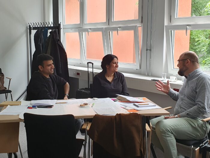 Three people from different countries sitting at the table filled with papers, by the window, talking and smiling. Troje ludzi z różnych krajów siedzących przy stole pełnym papierów, przy oknie rozmawiających i śmiejących się.