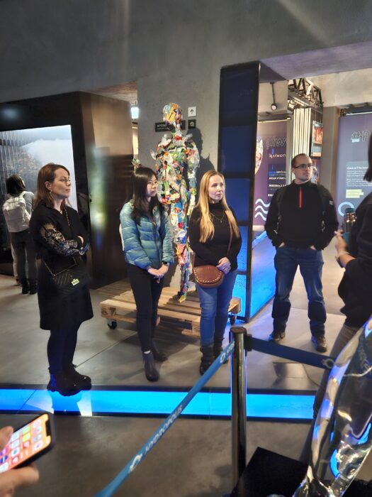 A group of people sitening in the water museum in the dim lights with a light stripe imitating riveer at their feet. Grupa słuchająca przewodniczki w muzeum, w przyciemnionym oświetleniu. Na podłodze widać niebieski pasek światła imitujący rzekę.