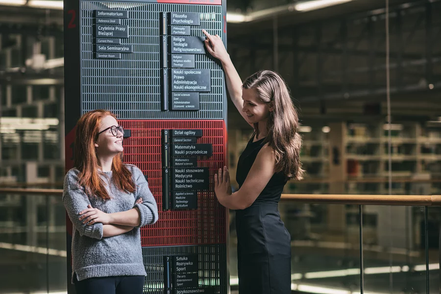 Dwie młode kobiety stojące przy tablicy informacyjnej z kierunkami studiów