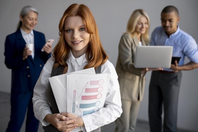 Młoda kobieta z teczkami w rękach, na dalszym planie osoba z telefonem komórkowym oraz dwie osoby spoglądające na ekran laptopa.