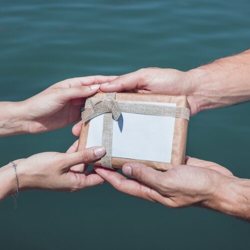 Dwie pary rąk trzymające prezent, w tle woda.