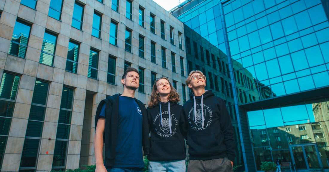 Trzy osoby w ubraniach z logo Uniwersytetu Śląskiego stoją na tle budynków i spoglądają w górę. Fot. Szymon Nawrat