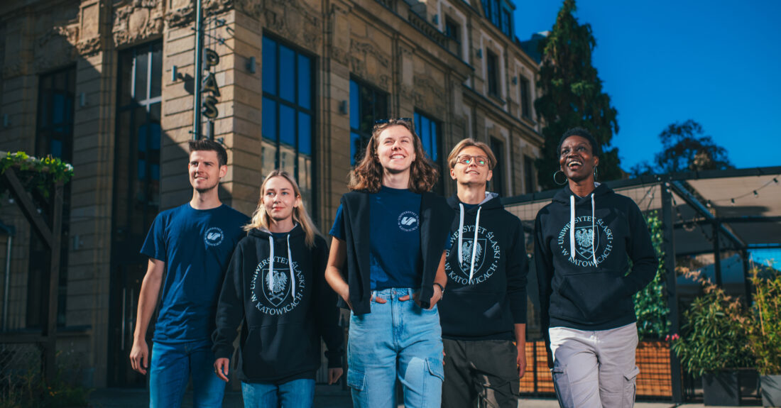Pięcioro studentów w odzieży z logo Uniwersytetu Śląskiego stoi na tle budynku.