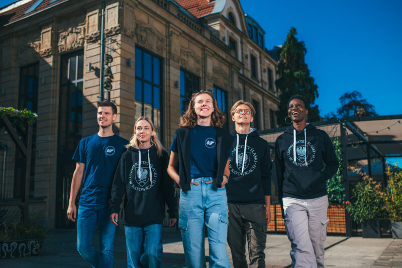 Pięcioro studentów w odzieży z logo Uniwersytetu Śląskiego stoi na tle budynku.