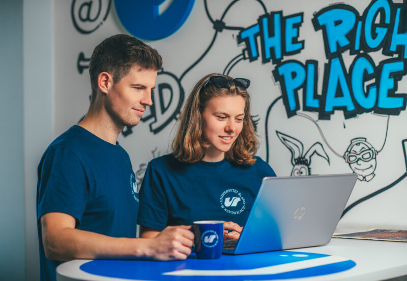 Dwoje studentów w koszulkach z logo Uniwersytetu Śląskiego stoi przy laptopie. Fot. Szymon Nawrat