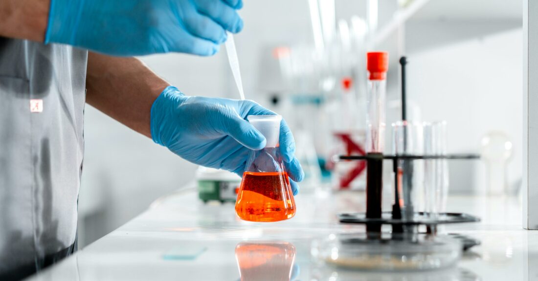 Osoba w rękawiczkach w laboratorium, trzymająca kolbę z płynem i pipetę.