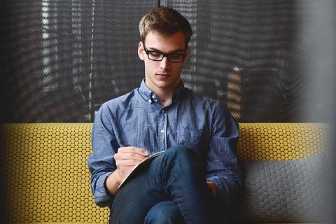 młody mężczyzna, w okularach - ubrany w koszulę i jeansy, siedzi na żółtej ławce i zaznacza coś na kartce