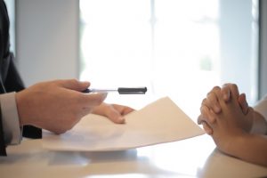 Zdjęcie przedstawia 2 osoby siedzące przy stole, przy czym jedna z nich trzyma długopis i kartkę papieru