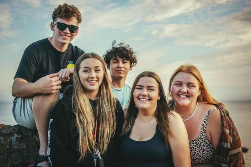 Zdjęcie przedstawia piątkę osób przyjaciół na tle zachodu słońca i wody. Wszyscy są uśmiechnięci