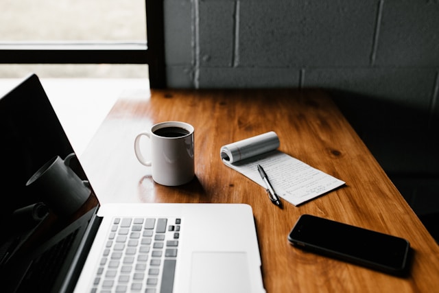 na biurku leżą: laptop, kubek z kawą, notatnik, długopis i telefon