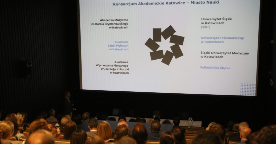 Zdjęcie przedstawia ludzi siedzących w auli wykładowej wpatrzonych w logo Europejskiego Miasta Nauki Katowice / People sitting at the lecture hall looking at the European City of Science Katowice logo