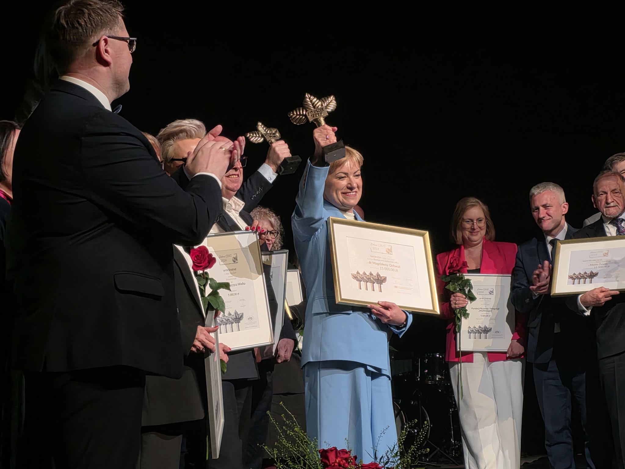 Na zdjęciu grupa ludzi kobieta trzyma statuetkę i dyplom w geście zwycięstwa
