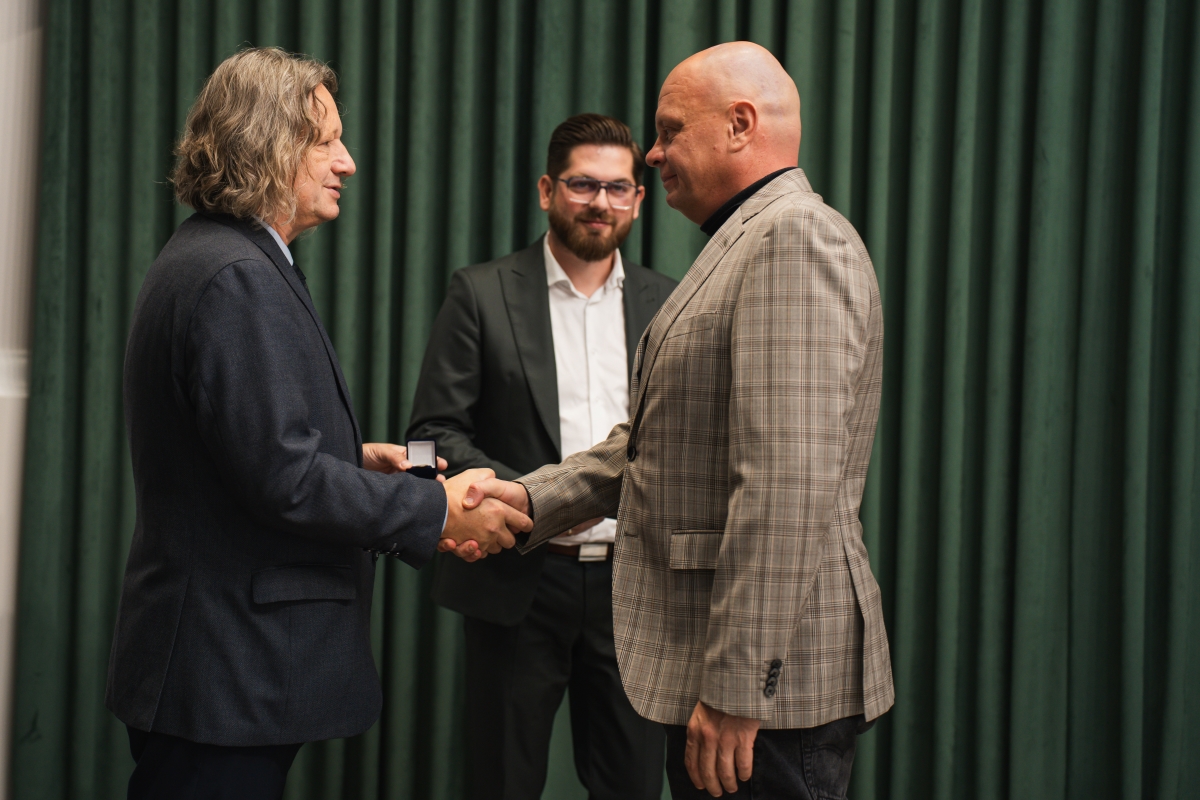 NA zdjęciu dwóch mężczyzn ściska swoje dłonie w tle widać uśmiechniętego mężczyznę