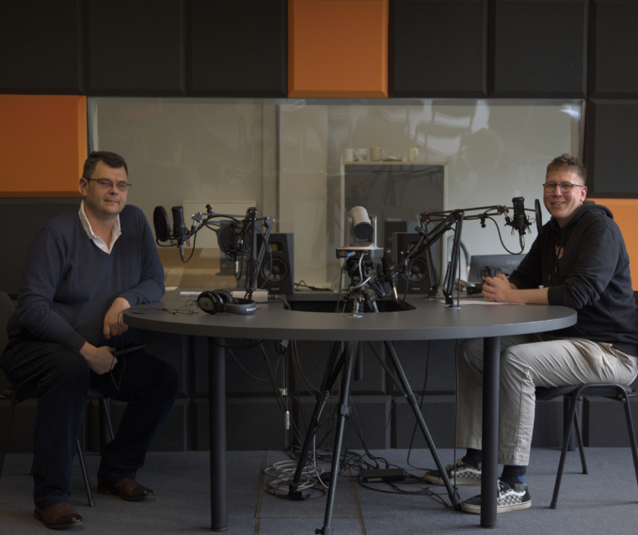 Zdjęcie przedstawia dwóch męzczyzn siedzących przy stole z mikrofonami w studiu podcastowym