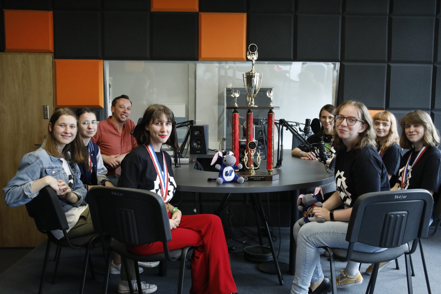 Na zdjęciu 6 uczestniczek konkursu wraz z trenerką oraz prowadzącym podcast. Siedzą w studiu radiowym na środku widać widowiskowy puchar i maskotkę usiołka