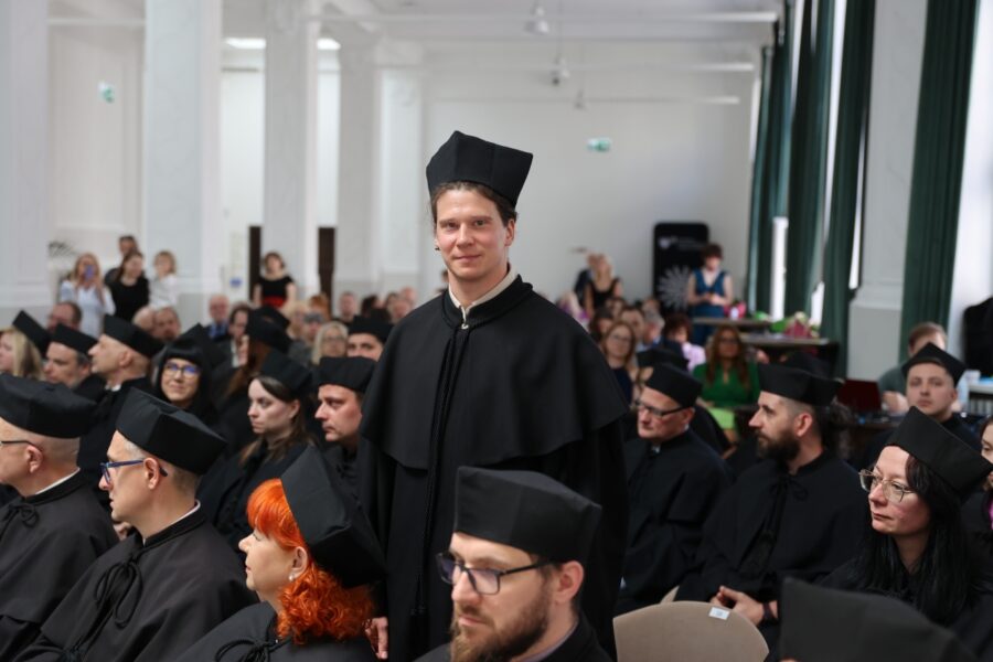 Uśmiechnięty doktorant pozuje do zdjęcie w koło siedzą doktoranci